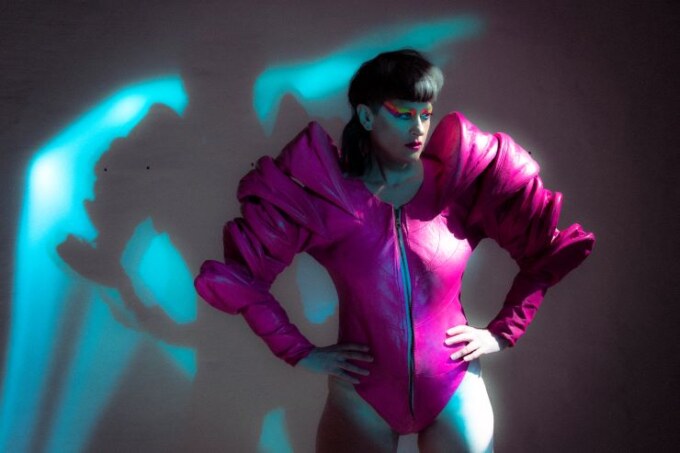 a press shot of the artist Peaches showing her in a dramatic pose with hands on hips wearing a pink leotard with structural shoulders and bold eye make up looking away from the camera